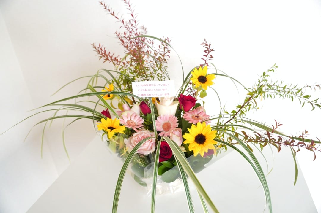 ひまわり、ピンクのガーベラ、赤いバラを緑の葉と薄い草でアクセントにしたフラワーアレンジメント。透明な花瓶に飾られています。